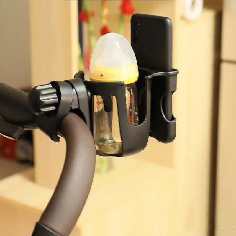 The stroller handle features a black bottle holder and phone holder attachment, versatile accessories part of the "Stroller Accessories" collection. These sturdy holders securely keep a baby bottle with a yellow cap and a smartphone in separate slots. In the background, you can see an indoor setting with blurred wooden furniture.