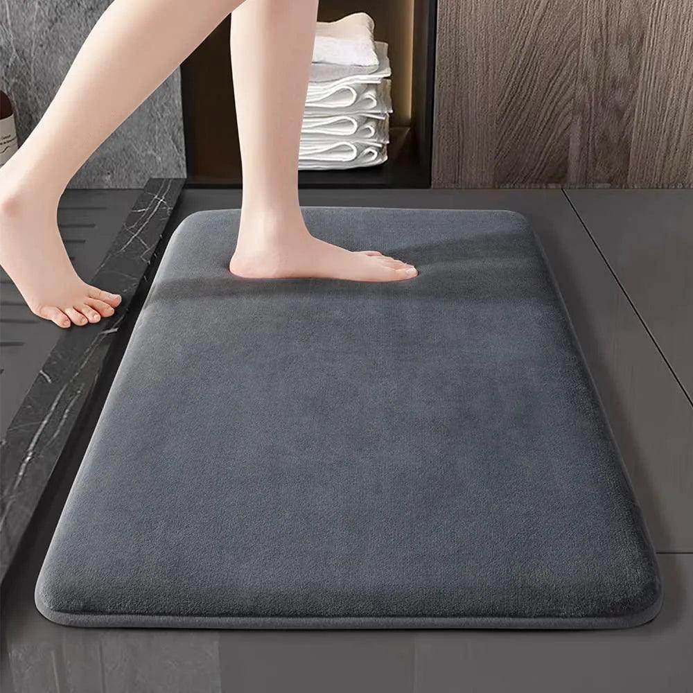 A person's bare foot steps onto a large-sized Super Absorbent Bath Mat, a Coral Velvet Floor Cover that is designed as an Anti-Slip Door Mat for the bathroom. In the background, neatly folded white towels are stacked on a shelf. The floor appears to be tiled, with a wooden panel nearby.