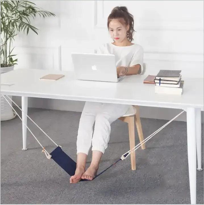 A person sits at a white desk using a laptop, enjoying the comfort of an "Under Desk Hammock." This portable folding office footrest pairs perfectly with the room's potted plant and stacked books, adding a charm that makes it an essential office accessory.