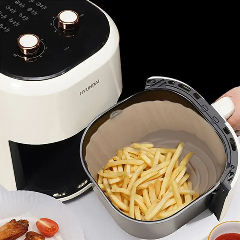 An OIMG brand air fryer with two dials is opened, showcasing a 1-piece reusable silicone basket filled with golden French fries. Beside it, a pie dish holds cooked meat and sliced cherry tomatoes against the stark black background.