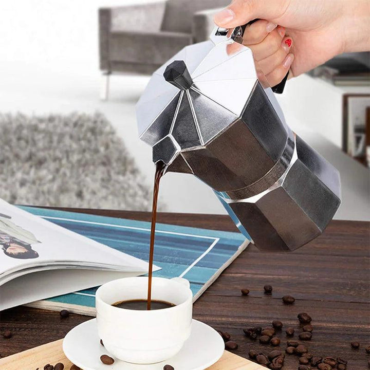 A hand pours coffee from the Moka Pot Espresso Maker, a Classic Italian Coffee Maker, into a white cup on a wooden table. Coffee beans are scattered around the table, and an open magazine is nearby. A gray sofa and rug are visible in the blurred background.