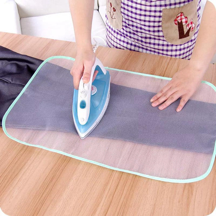 A person is carefully ironing a piece of dark polyester fiber on a wooden table with a blue and white iron. They use the Protective Press Mesh, Insulation Ironing Board Mat to shield the fabric, while wearing a checkered apron with pockets. Nearby, plastic storage boxes keep everything organized.