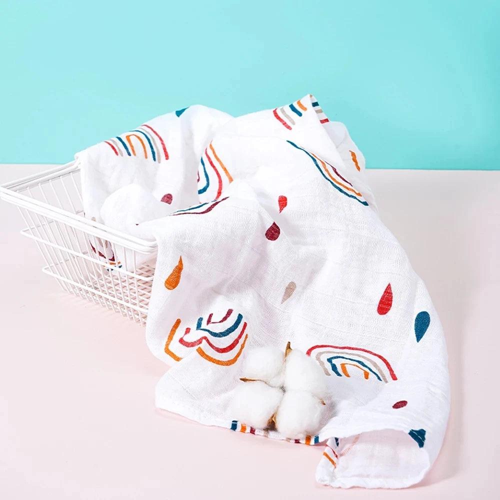 A vibrant Baby Bib Burp Cloth Set Bamboo Cotton Muslin Bib, adorned with rainbow and raindrop designs, elegantly covers a white wire basket. Three cotton bolls rest atop the cloth, set against a pastel blue wall and light pink surface—ideal for showcasing any baby essentials gift set.