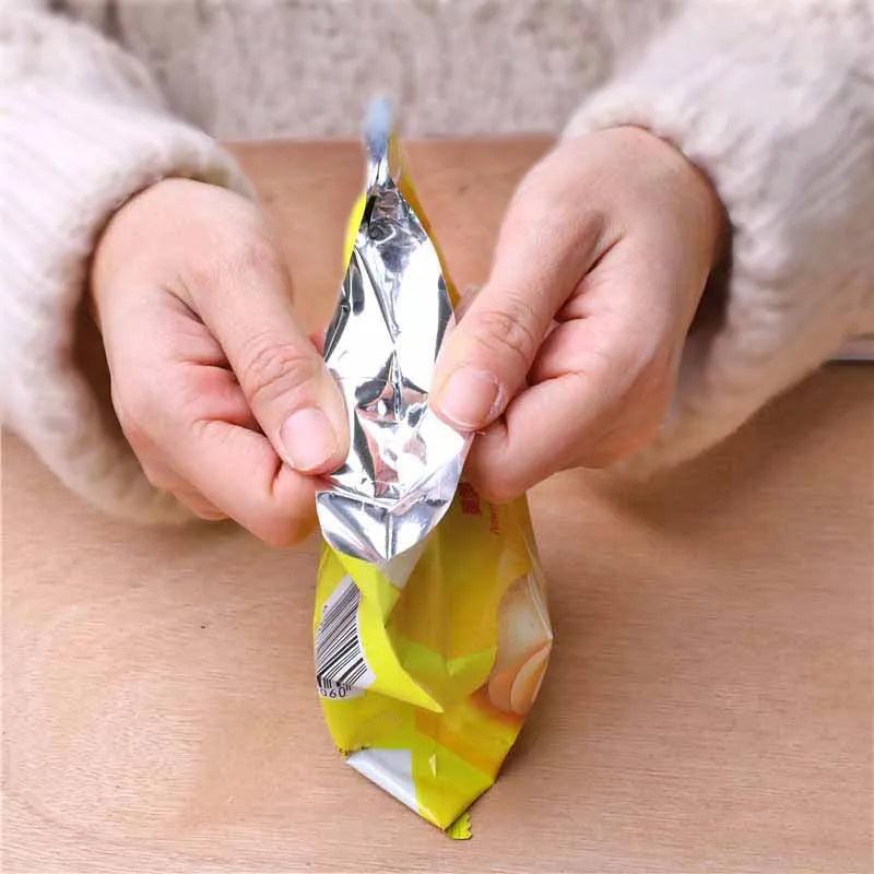 Hands holding an unopened yellow snack bag with a shiny silver interior, resting on a wooden surface, ready to be sealed using the "Heat Bag Sealer, Food Packaging Sealing Machine, Portable Snack Bag, Sealing Clip.