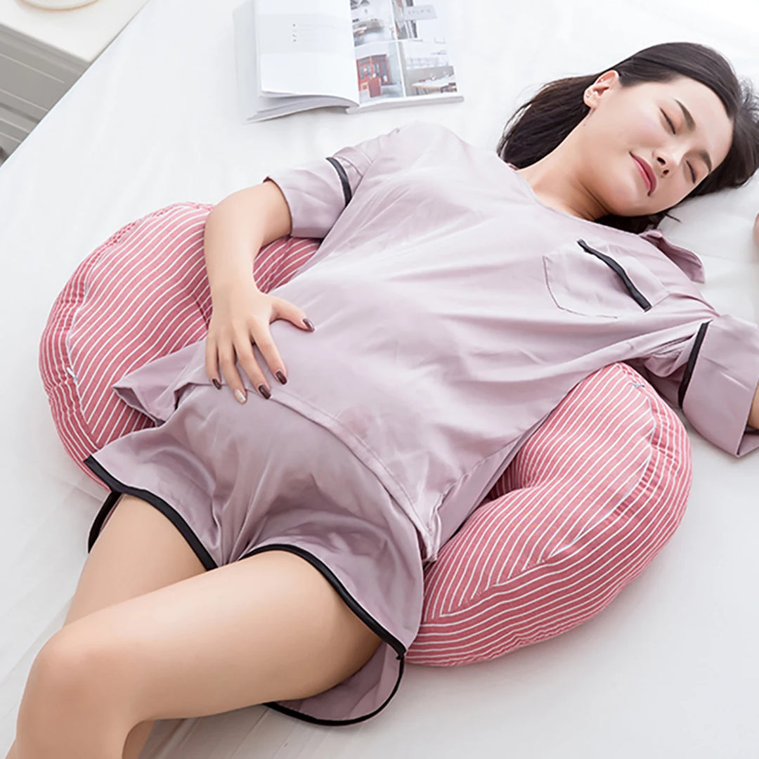 A pregnant woman in pajamas lies comfortably on a bed, supported by a Pregnancy Pillow, Side Sleeping Pillow, and U-Shaped Maternity Pillow that cradles her back and belly. She appears relaxed with one arm resting on her stomach.