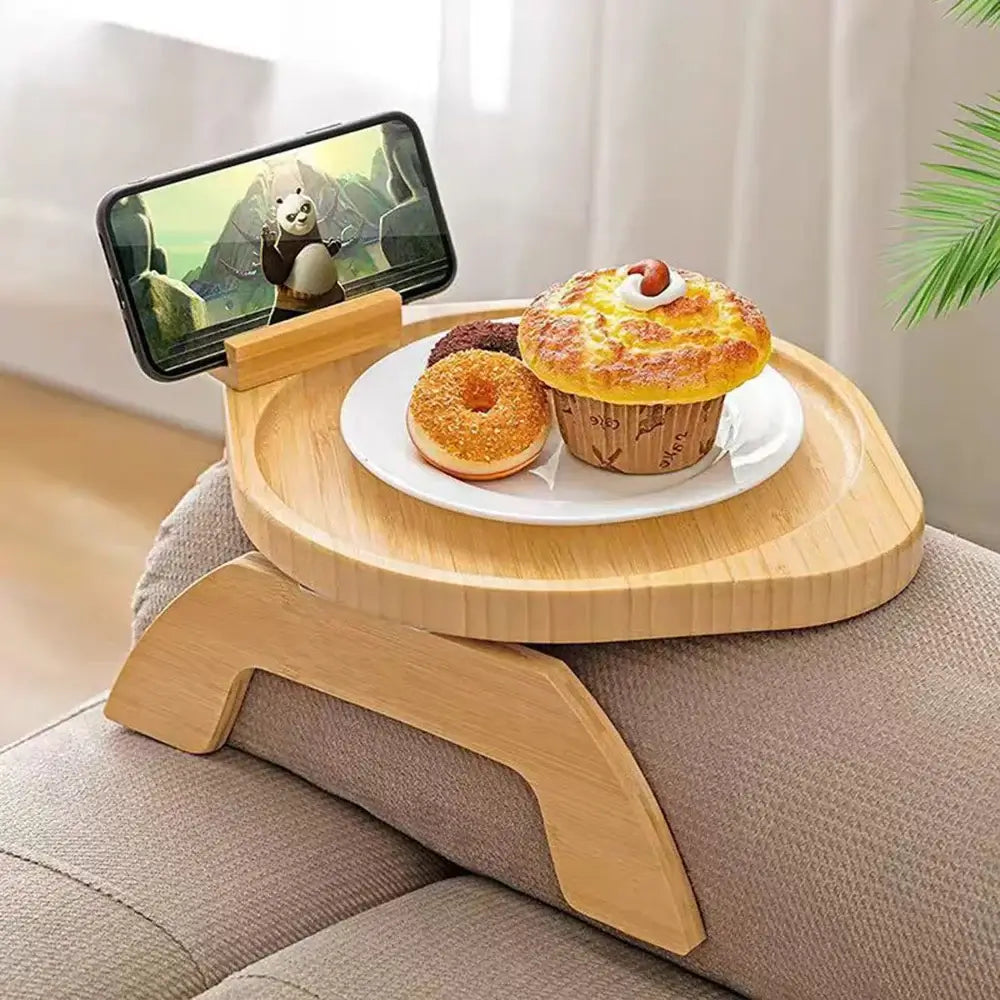 A Tray for Sofa, Tray Table sits on a sofa armrest, holding a smartphone with a panda animation playing. This adaptable tray also accommodates a plate with a muffin, donut, and cookie. In the background, there's a lush palm plant.