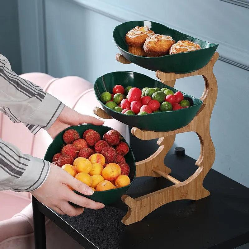 A wooden tableware set, crafted from eco-friendly materials, holds green bowls filled with pastries, mixed grape tomatoes, and a blend of strawberries and yellow plums. A person is placing the bottom bowl on the kitchen serving trays. The setup is on a black table near a pink chair.