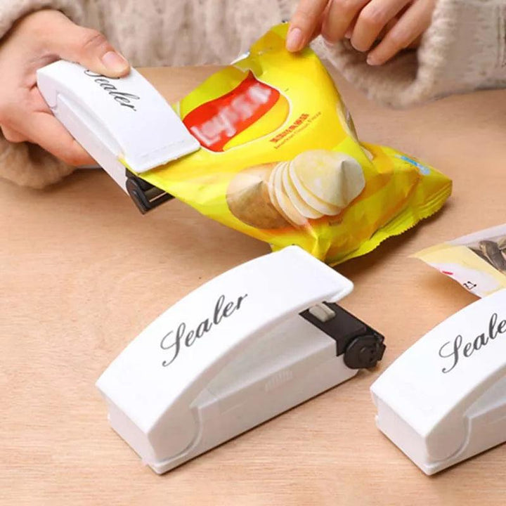 A person is using a white "Heat Bag Sealer, Food Packaging Sealing Machine, Portable Snack Bag, Sealing Clip" to secure a yellow potato chip bag on a wooden surface, ensuring the snacks stay fresh. Two additional sealers are on the table along with handy sealing clips for convenient food storage.