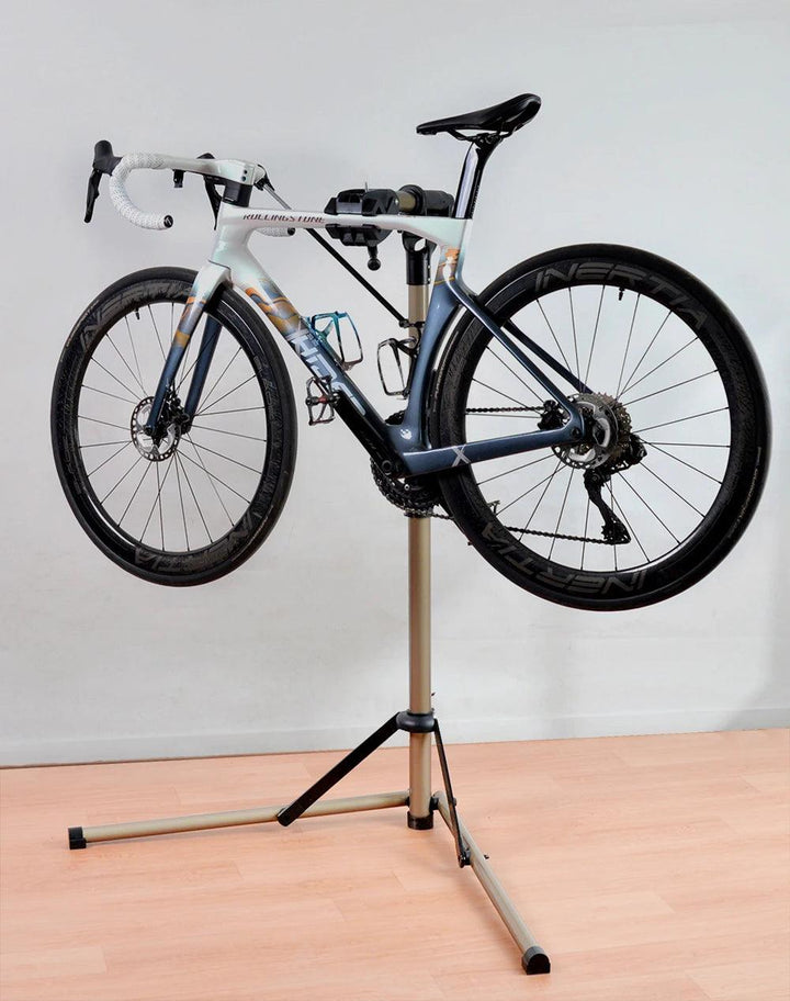 A road bicycle with drop handlebars is gracefully positioned on the Bike Repair Stand Professional Bike Maintenance Foldable Bicycle Repair Stand. The bike, featuring a white and blue frame paired with black wheels, is set against a plain light-colored wall and wooden floor, perfectly prepared for any maintenance it might need.