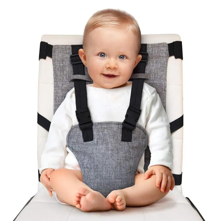 A baby with light hair and blue eyes sits smiling in a gray harness attached to a white dining chair, using the Child Chair Safety Belt Adjustable Kids Feeding Belt Baby Dining Chair Seat Belt. The adjustable safety belt keeps the baby secure, while the water-resistant materials ensure easy cleanup. The little one wears a white outfit and sits with crossed legs.