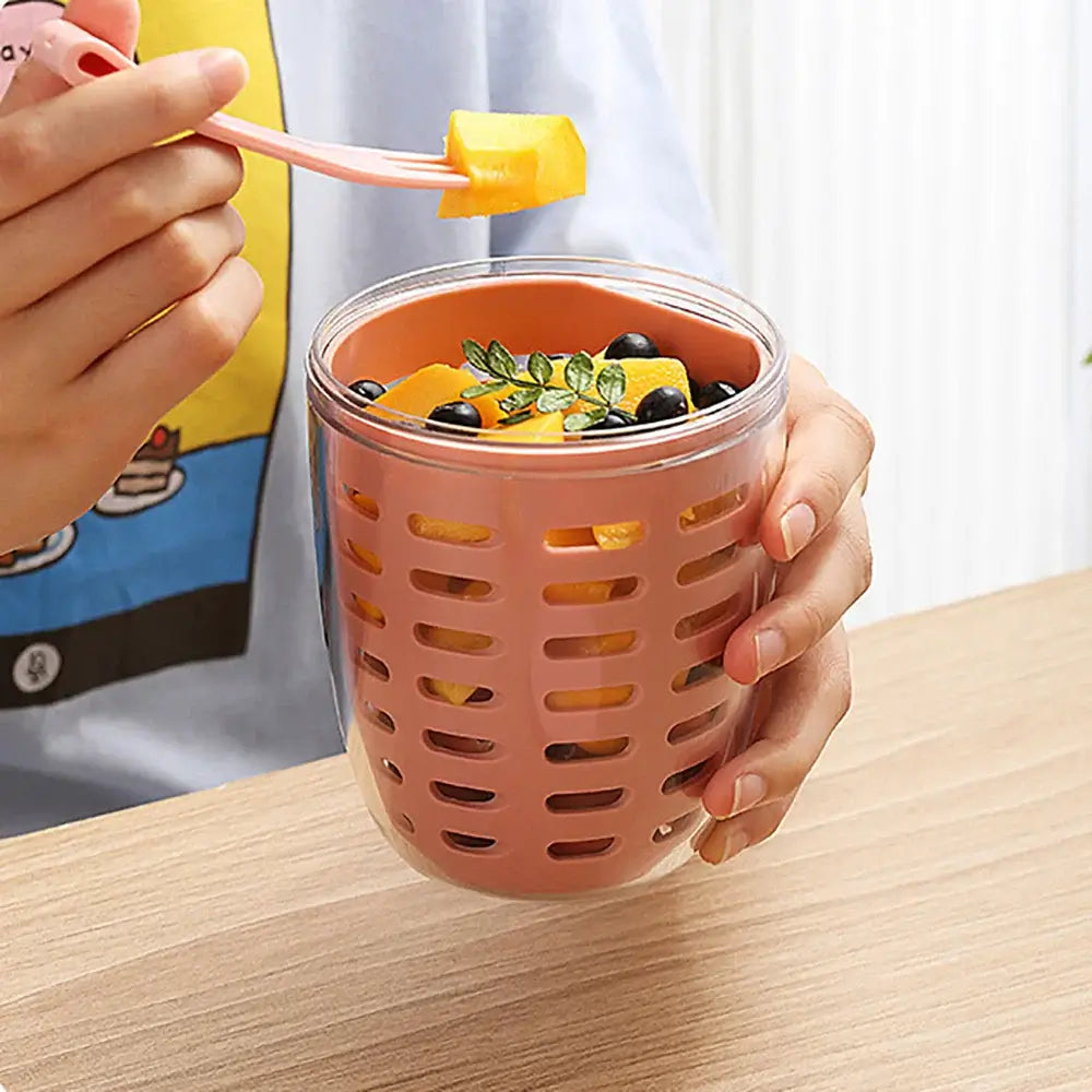 A person is holding the "Fruit Cup with Fork Portable Picnic Bottle," an eco-friendly, pink lattice-designed container filled with diced mangoes, blueberries, and a small green leaf garnish. Using the included fork, they're taking a piece of mango while the cup sits securely without spills on a wooden surface, making it perfect for use as a fruit strainer bowl for any occasion.