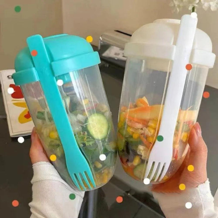 Two hands hold clear Portable Bottle Salad Cups, with vibrant salads inside. One of the containers serves as a comprehensive lunch box, featuring a teal lid and fork, while the other showcases a white set. Made from food-grade PP material, they are placed against an abstract decorative backdrop on the table.