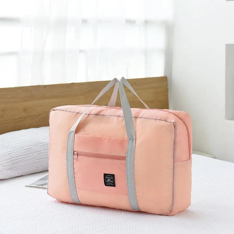 The Large Capacity Foldable Travel Bag in peach, featuring gray handles and a front zipper pocket, rests on a white bed. The background showcases a wooden headboard and sheer white curtains, highlighting the bag's waterproof design ideal for convenient travel.