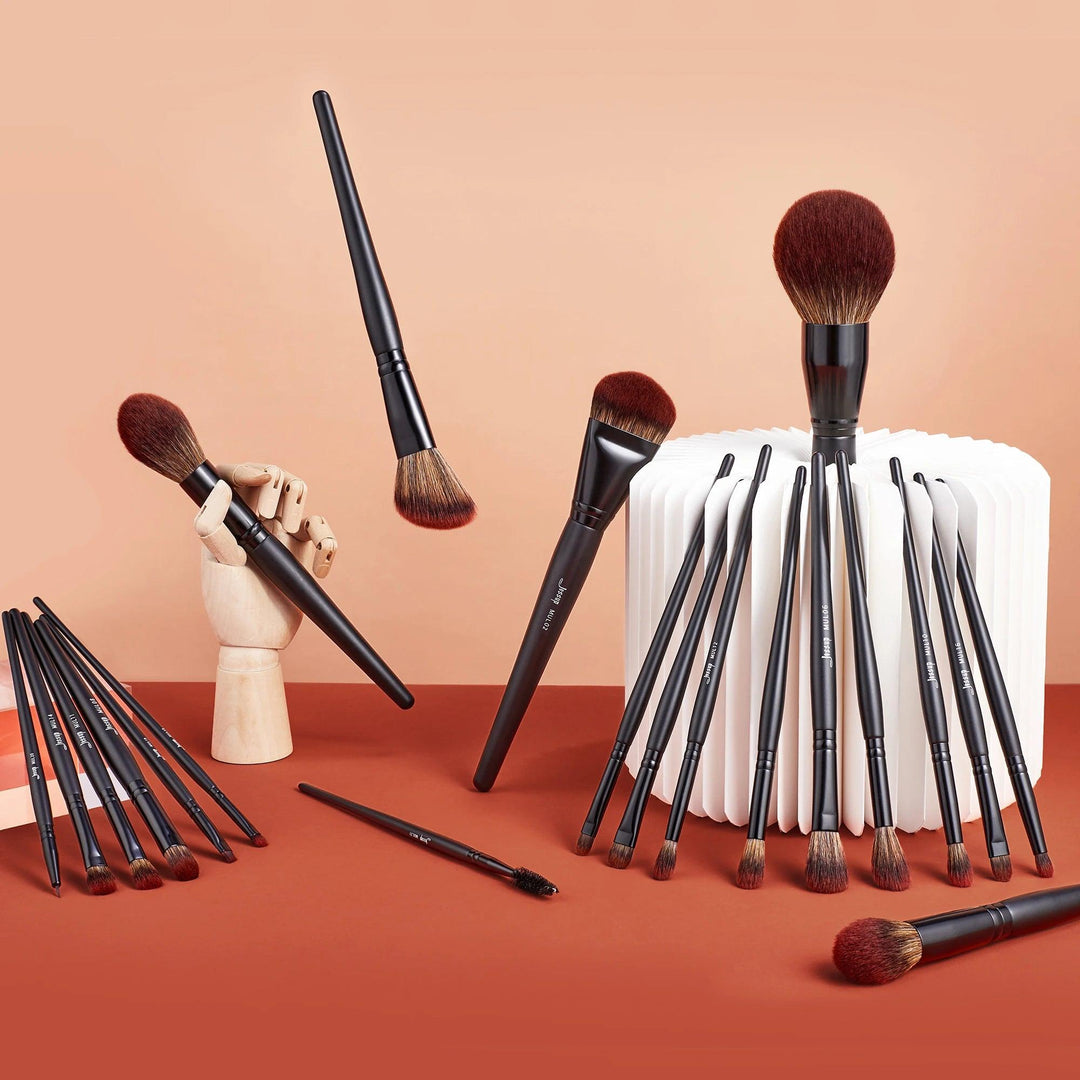 A collection of the Makeup Brushes Set featuring synthetic powder, concealer, eyeshadow, and eyeliner tools with dark handles and red-brown bristles is displayed on an orange surface. Some brushes are propped up creatively, including one positioned in a wooden hand model. A white textured surface serves as the background against a peach wall.