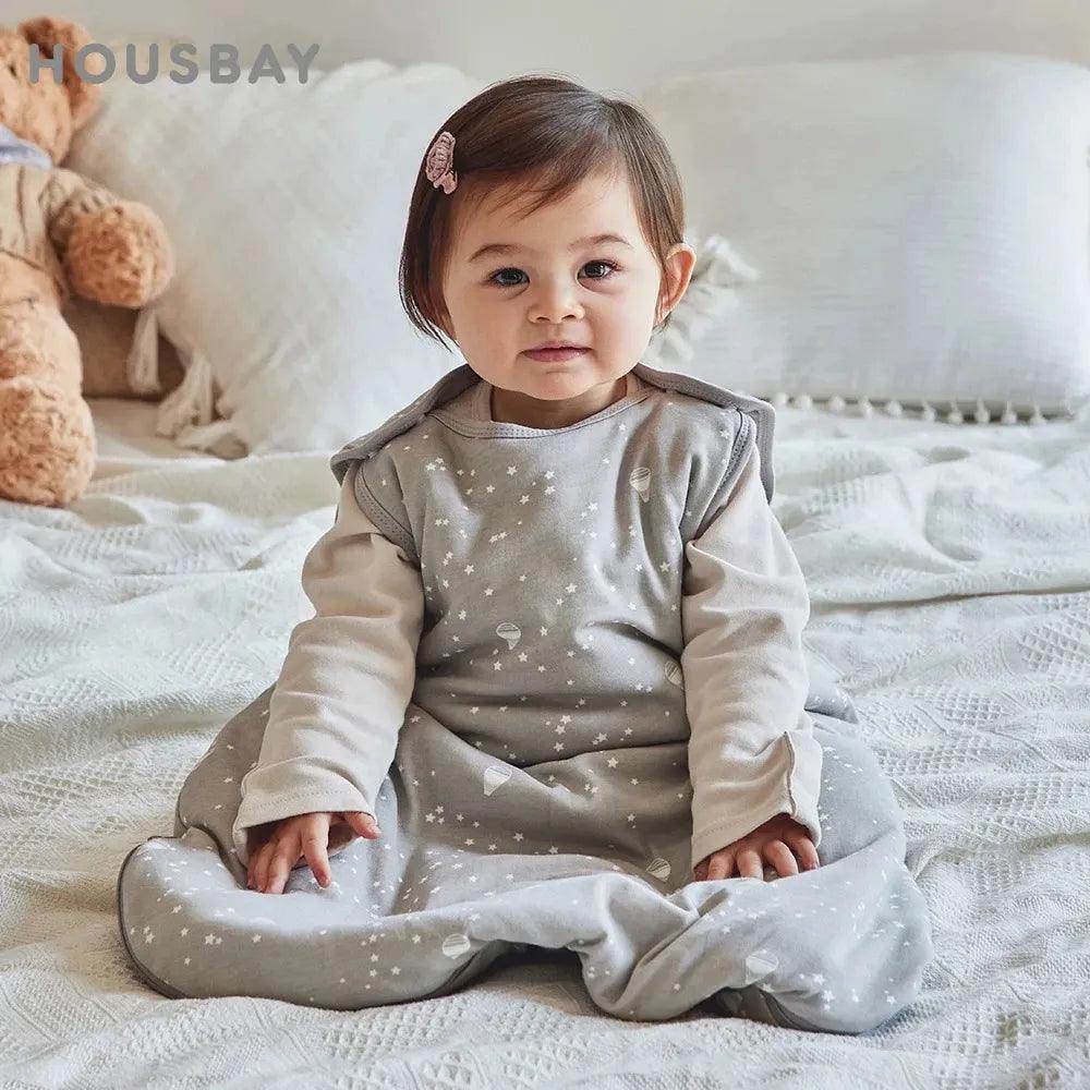 A baby sits on a bed in the Baby Sleep Sack, designed for all seasons with a gray star pattern and crafted from cozy cotton polyester. Wearing a long-sleeved shirt and a small hair clip, the little one is surrounded by soft pillows and a teddy bear in the background.