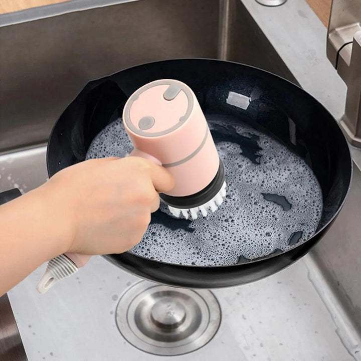 A person uses a pink Cordless Electric Bathroom, Kitchen & Sofa Cleaning Brush equipped with a built-in soap dispenser to scrub a frying pan with soapy water in the kitchen sink.