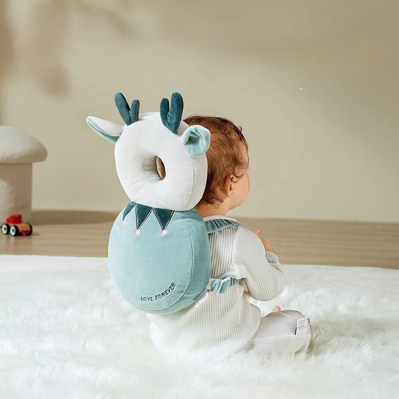 A baby is sitting on a soft white rug, dressed in a light-colored onesie and wearing an adorable backpack shaped like a deer with antlers and ears. A Baby Head Protection Infant Safety Headrest Cushion adds extra protection, while a small toy car lies on the wooden floor in the background.