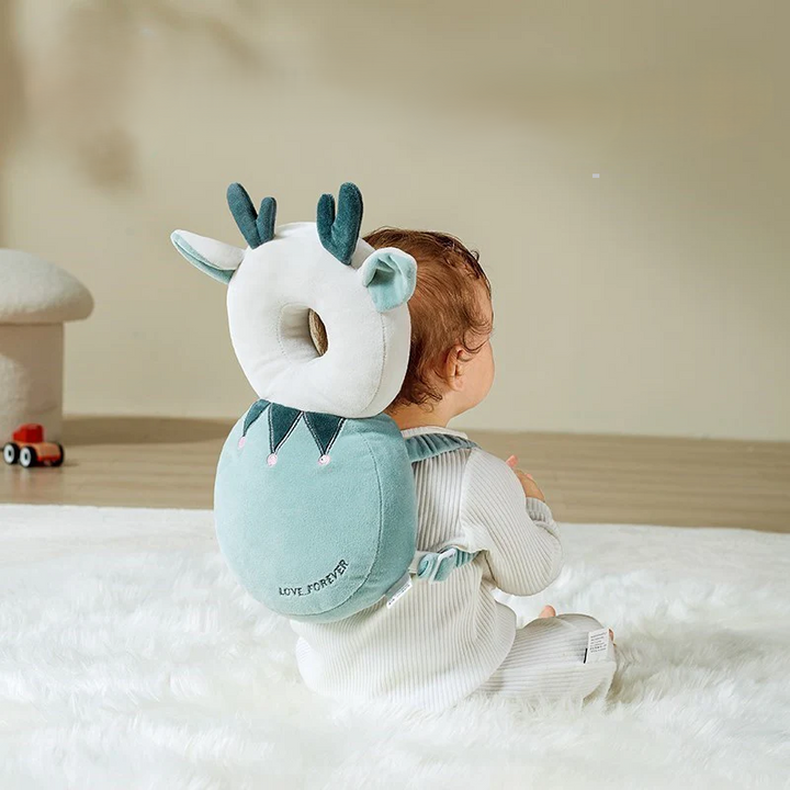 A baby is sitting on a soft white rug, dressed in a light-colored onesie and wearing an adorable backpack shaped like a deer with antlers and ears. A Baby Head Protection Infant Safety Headrest Cushion adds extra protection, while a small toy car lies on the wooden floor in the background.