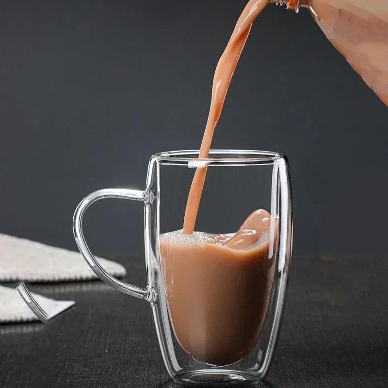 A Double-Layer Transparent Cup made of high borosilicate glass, designed to be heat resistant, is being filled with creamy brown tea from a bottle. The cup is set on a dark, minimalist surface, against a backdrop of plain dark grey.