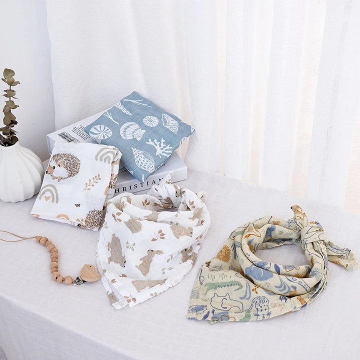 A minimalist scene features two patterned cotton drool bibs, part of the Muslin Blanket Set Baby Drool Bibs Cotton Burp Cloth, displayed on a light-colored tablecloth. A pacifier clip adorned with wooden beads is placed next to them. In the background, books and decorative cushions showcasing hedgehog and seashell designs are arranged alongside a soft muslin blanket.