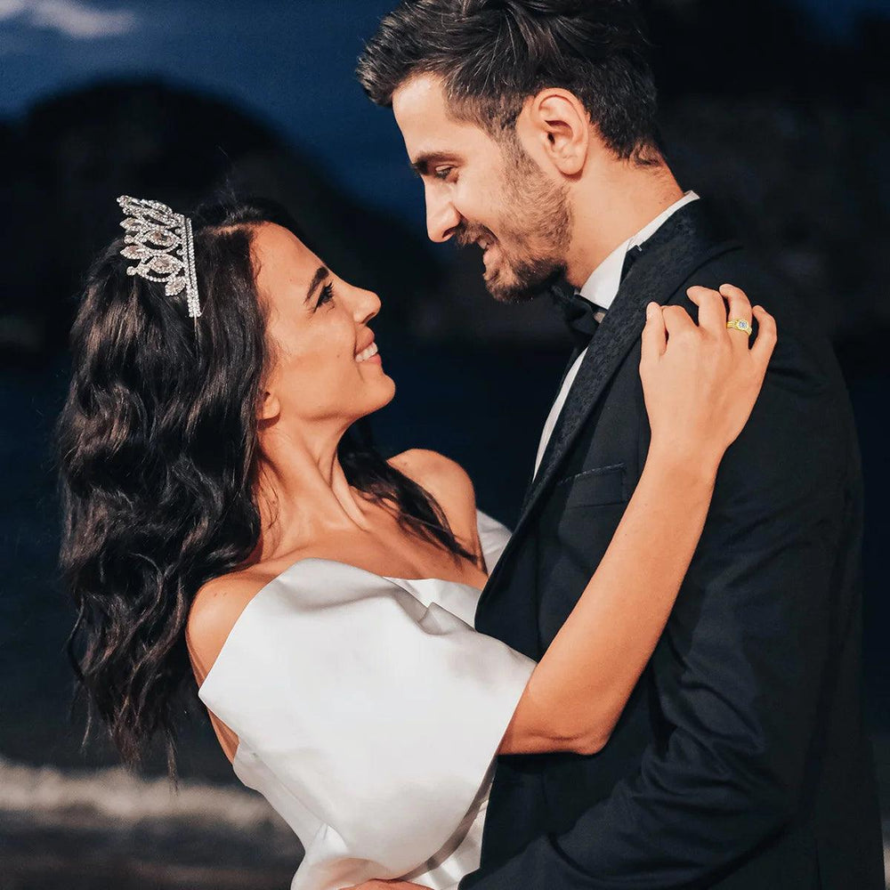 A couple dressed in formal attire embraces, smiling against a dark outdoor backdrop. The woman, adorned with a tiara and white dress, showcases a Moissanite Engagement Ring from the Bow Knot Wedding Band collection, while the man stands beside her in a black suit.