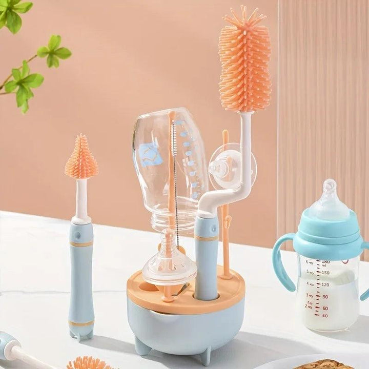 The Portable Baby Bottle Cleaner Set, made with durable plastic, includes orange silicone brushes and blue handles, designed to securely hold an upside-down baby bottle for drying. A sippy cup and pacifier are placed on a white surface nearby with a green-leaved plant enhancing the natural ambiance in the background.