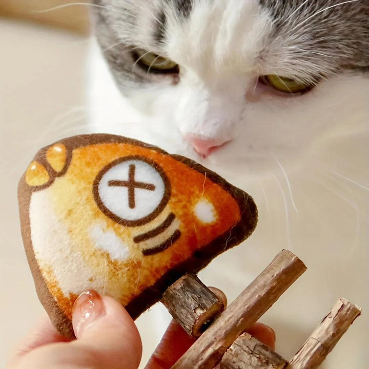 A cat with white and gray fur sniffs a plush roast fish toy for cats, designed like an orange fishbone with large eyes and an X for a mouth. The toy, potentially infused with enticing catnip, is held by a hand using a wooden clip.