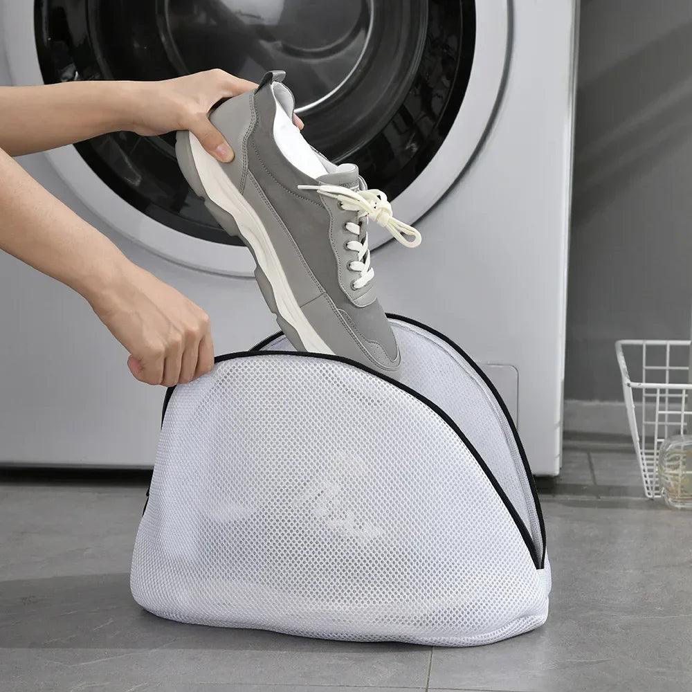 A person places a gray sneaker into a white Mesh Laundry Bags Shoes Storage Organizers Travel Shoes Storage Bag before loading it into the washing machine, while a laundry basket sits on the floor nearby.