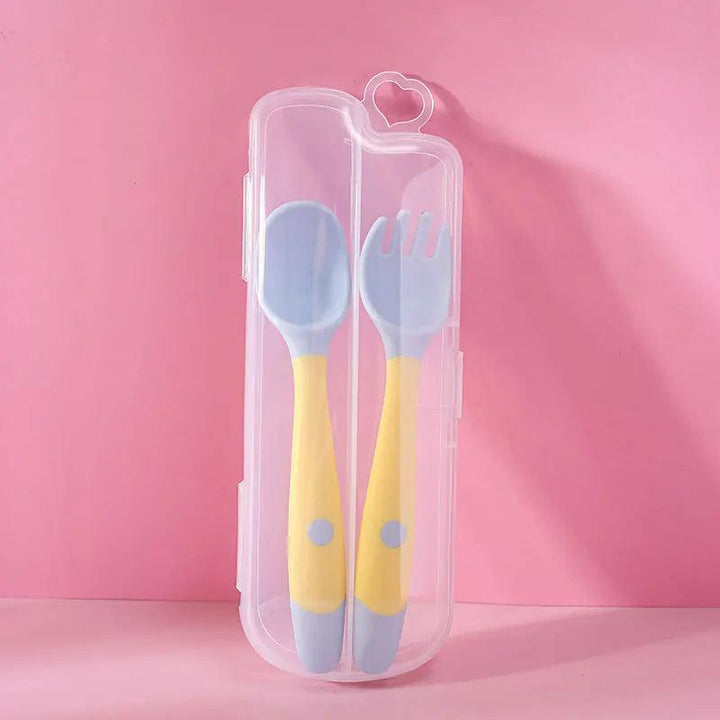 A pastel blue and yellow Baby Feeding Spoon Training Utensils set, adorned with polka dots and made from BPA-free silicone, includes a spoon and fork designed with non-slip grips for little hands. The set is stored in a clear case featuring a heart-shaped clasp, all displayed against a pink background.