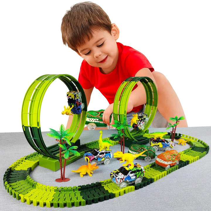 A young boy in a red shirt is captivated by the Dinosaur Car Track Set, Flexible Race Track, Toy Kid's Toy Car Set. This educational toy includes two large green loops where battery-operated toy cars race past small dinosaur figures and palm trees on a gray surface.