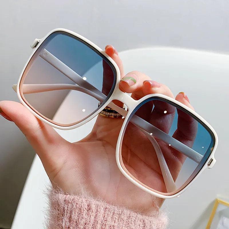 A hand holds the Oversized Rectangle Sunglasses Women's Fashion Sunglasses, featuring white frames and dark lenses that elegantly reflect light. Paired with a pink, fuzzy sweater, they showcase a perfect blend of fashion-forward women's eyewear.