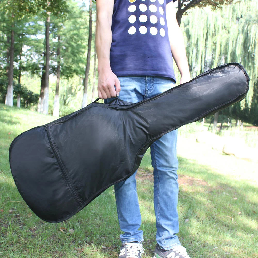 A person wearing blue jeans and a navy shirt stands outdoors in a park, surrounded by green grass and trees, holding the Portable Acoustic Guitar Bag—a soft case with a shoulder strap from the Guitar Parts & Accessories collection.