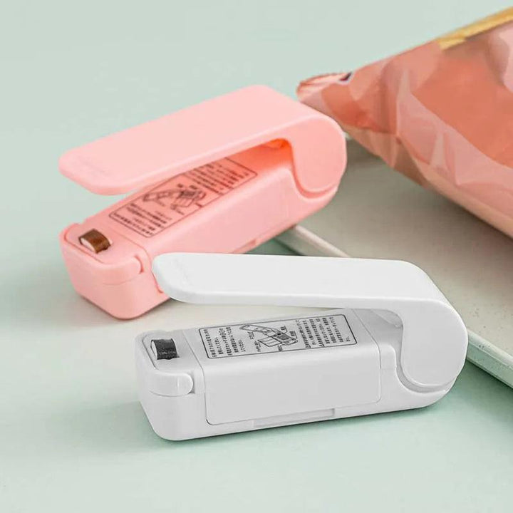 A pink and a white version of the "Heat Bag Sealer, Food Packaging Sealing Machine, Portable Snack Bag, Sealing Clip" rest on a light green surface, ideal for sealing food storage bags. These sealers feature flip-up lids that reveal metal heating strips. In the background lies a partially visible pink bag, ready to benefit from this convenient food-saving tool.