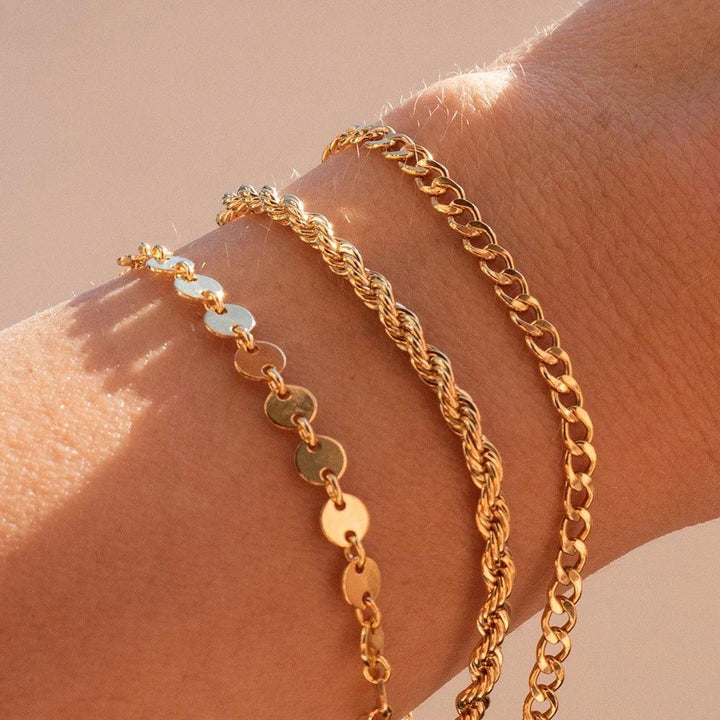 A close-up shot features a wrist elegantly decorated with three distinct gold bracelets: the "Trendy Cuban chain bracelet," the stylish "Snake chain bracelet," and the sophisticated "Gold plated stainless steel bracelet" adorned with small round discs. The softly blurred background accentuates their captivating allure.