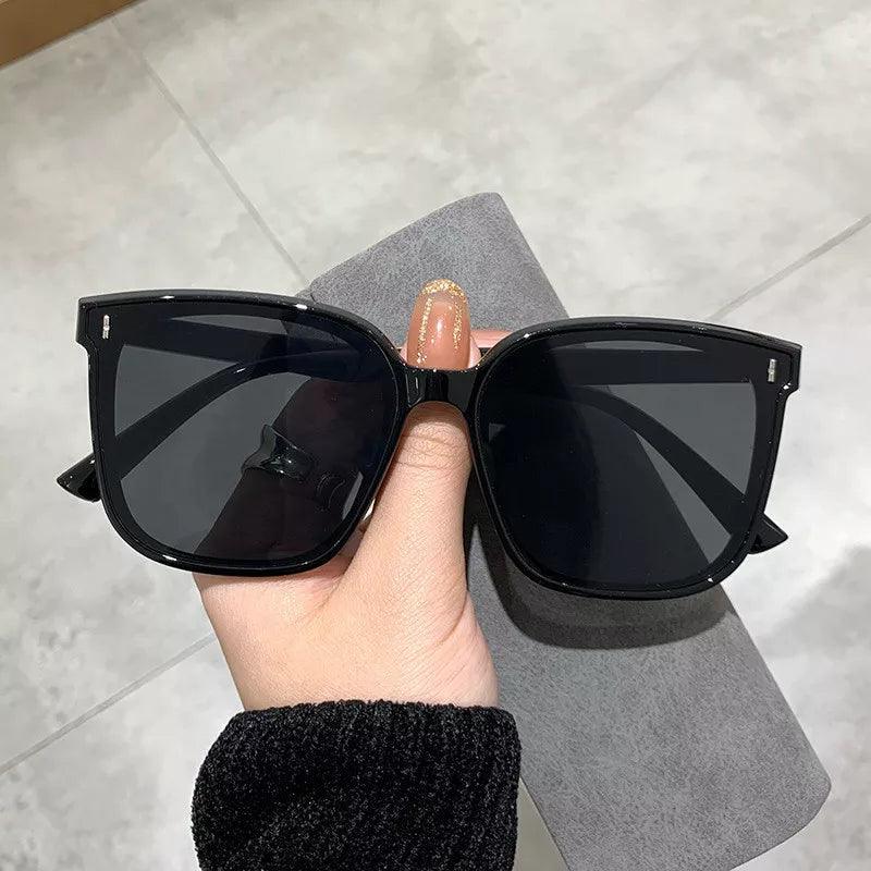 Against a light gray background, an individual with painted nails and a dark sweater holds "Oversized retro shades," featuring black polished frames and dark tint. The "Square sunglasses" offer UV400 protection, and the person also has a soft cloth in their hand.