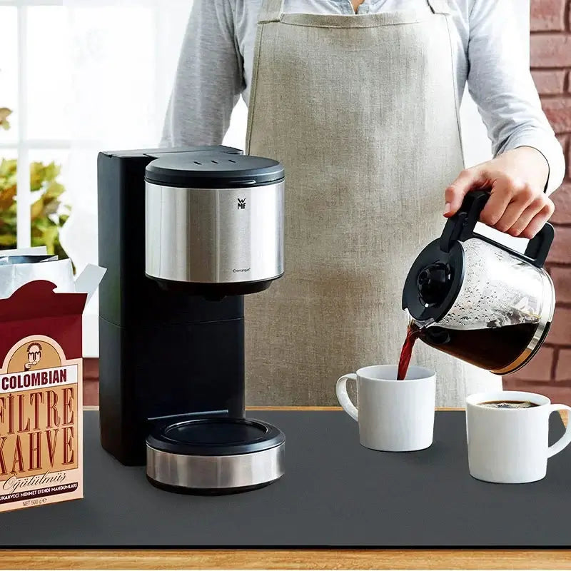 Person pouring coffee into mugs beside coffee machine on kitchen mat.