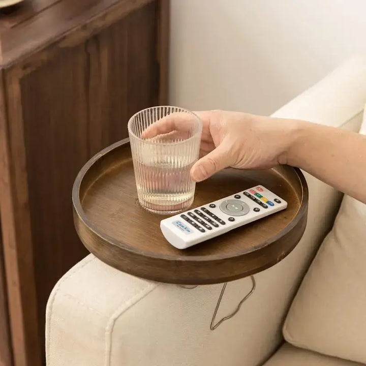 A hand holding a glass of water on the Sofa Armrest Table, featuring a side wooden storage tray for snacks, tea, or coffee attached to a beige sofa. A remote control also rests on this multifunctional round tray, with a wooden cabinet visible in the background.