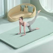 Wearing a light-colored outfit, a person is exercising on a 10mm thick yoga mat made of high-quality TPE with an anti-slip design in a modern room. They are in a kneeling pose, lifting one leg. Behind them is a curved beige sofa adorned with several cushions, while large windows fill the space with natural light.